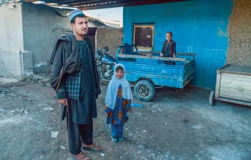 Sahib Chan stoi przed domem ze swoimi dziećmi. Za nim motorynka, dzięki której zarabia na życie. Czarahi Kambar, luty 2021 r. / PAWEŁ PIENIĄŻEK