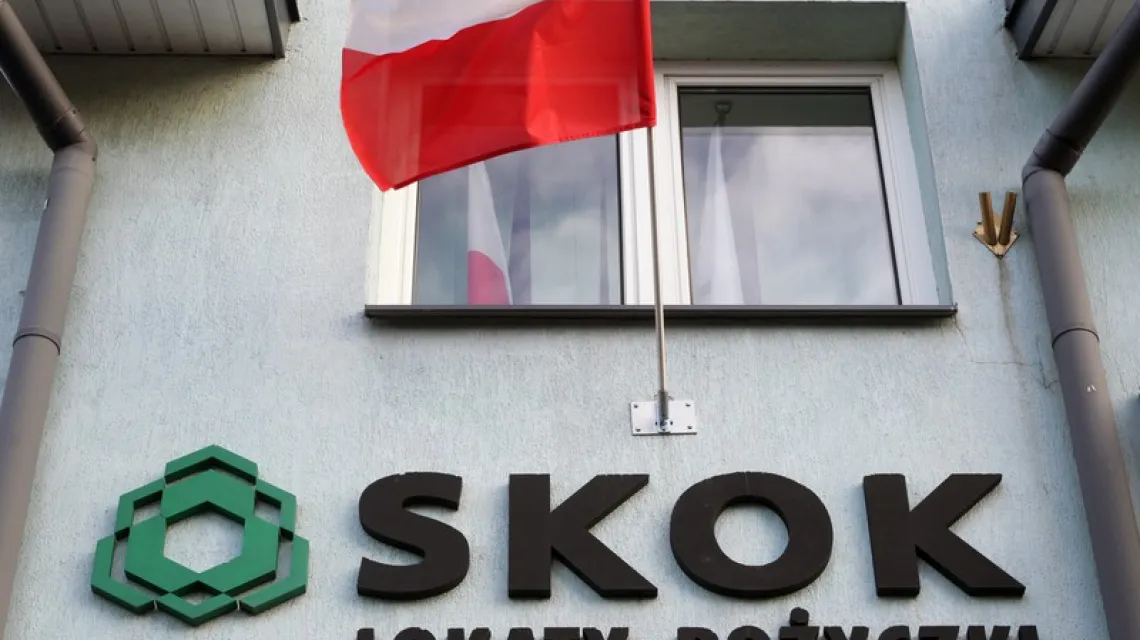 Logo SKOK pod oknem, nad którym powiewa flaga biało-czerwona