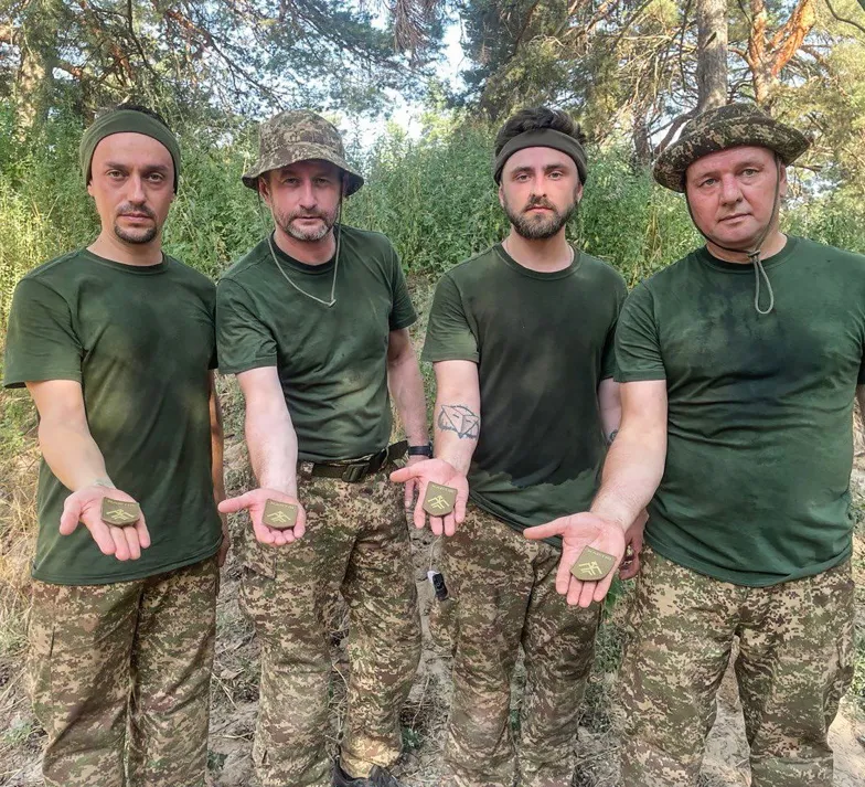 Serhij Żadan (2. z lewej) z kolegami z wojskowego szkolenia demonstrują naszywki brygady „Chartia”. 1. z prawej Ołeksandr Merenczuk, puzonista zespołu Żadan i Sobaki, który wstąpił do wojska razem z Serhijem, opowiadał, że naszywki „Chartii” nie da się kupić w żadnym ze sklepów militarnych (w odróżnieniu od emblematów innych jednostek); by ją zdobyć, nieźle się zadyszał podczas ostatniego egzaminu na poligonie: kilkukilometrowego biegu w pełnym uzbrojeniu. Wiosna-lato 2024 r. // Fot. archiwum prywatne