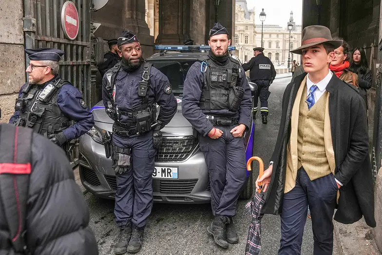 Po kradzieży klejnotów z Luwru fotograf Thibault Camus uchwycił na zdjęciu elegancko ubranego mężczyznę przechodzącego obok policjantów blokujących wejście do muzeum. Obraz obiegł świat, stając się internetową sensacją. Użytkownicy mediów społecznościowych okrzyknęli nieznajomego detektywem, choć Camus twierdzi, że to po prostu przypadkowy przechodzień. A może francuska policja trzyma jego personalia w tajemnicy? Paryż, 19 października, 2025 r. // Fot. Thibault Camus / AP / East News 